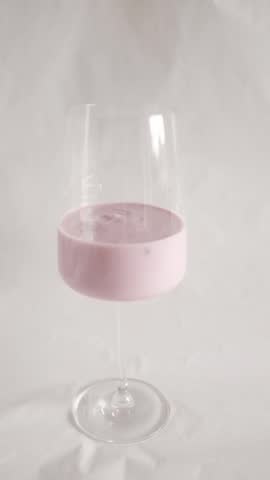A vertical, clean studio shot of creamy pink berry yogurt served in an elegant, long-stemmed wine glass against a seamless white background. The video features a smooth, slow zoom-out that starts close on the swirling, glossy texture of the yogurt—revealing hints of blended fruit pieces—and moves back to showcase the minimalist, sophisticated presentation of the entire glass. The bright, soft, diffused lighting highlights a healthy, aesthetic, and appetizing snack concept. Ideal for dairy brand commercials, healthy lifestyle vlogs, breakfast menus, and gourmet food styling content.