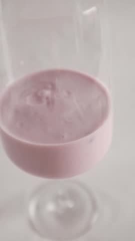 A vertical, clean studio shot of creamy pink berry yogurt served in an elegant, long-stemmed wine glass against a seamless white background. The video features a smooth, slow zoom-out that starts close on the swirling, glossy texture of the yogurt—revealing hints of blended fruit pieces—and moves back to showcase the minimalist, sophisticated presentation of the entire glass. The bright, soft, diffused lighting highlights a healthy, aesthetic, and appetizing snack concept. Ideal for dairy brand commercials, healthy lifestyle vlogs, breakfast menus, and gourmet food styling content.