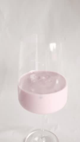 A vertical, clean studio shot of creamy pink berry yogurt served in an elegant, long-stemmed wine glass against a seamless white background. The video features a smooth, slow zoom-out that starts close on the swirling, glossy texture of the yogurt—revealing hints of blended fruit pieces—and moves back to showcase the minimalist, sophisticated presentation of the entire glass. The bright, soft, diffused lighting highlights a healthy, aesthetic, and appetizing snack concept. Ideal for dairy brand commercials, healthy lifestyle vlogs, breakfast menus, and gourmet food styling content.
