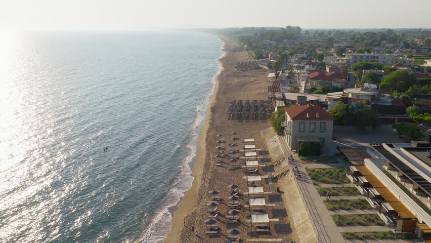 Drone flies over sandy Kourouta beach, Peloponnese, Greece. Cafe-bar beach umbrellas, bathers on shore. Clear sunny summer day