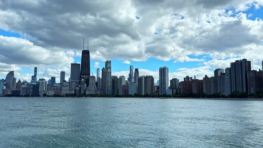 Tall buildings of Chicago line the lake shore with a clear view of the skyline. The water reflects the clouds and blue sky above. Many people enjoy the scenic view nearby.