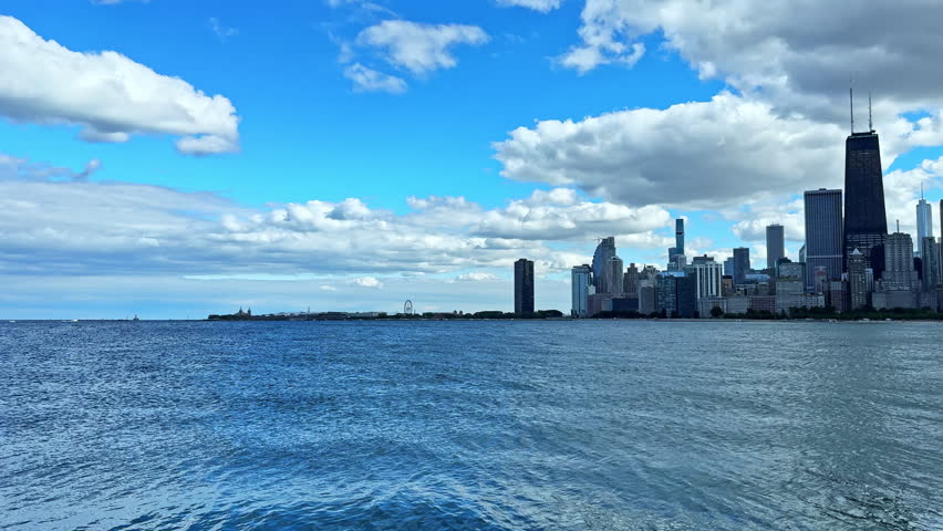 People can see the Chicago skyline while standing near the water. The blue sky has clouds and the lake reflects the city. Buildings are visible along the shoreline.