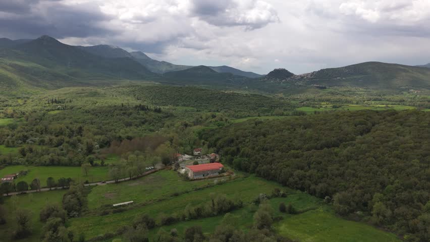 Wide aerial drone footage of a lush green mountain valley in the Peloponnese, Greece. The landscape features olive groves, forested hills, open fields, scattered small farm buildings, distant hillside houses and layered mountain ranges under dramatic clouds. Soft sunlight breaks through the cloud cover and creates moving patches of light and shadow across the spring greenery. This footage is suitable for nature, travel, scenic landscape, Mediterranean scenery, mountain views, peaceful outdoor backgrounds, aerial establishing shots, environmental visuals and southern European landscape projects.