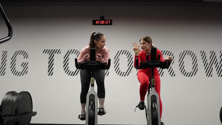 Two women on stationary bikes count down before friendly fitness race pushing speed endurance and competitive spirit during intense cardio training session