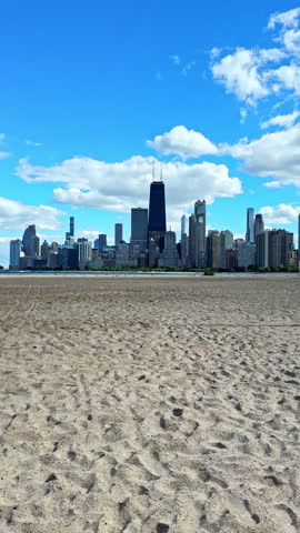 People enjoy a sunny day at the beach while the Chicago skyline rises in the background. The sandy area is vast and the sky has some clouds. The buildings stand tall against the blue sky.