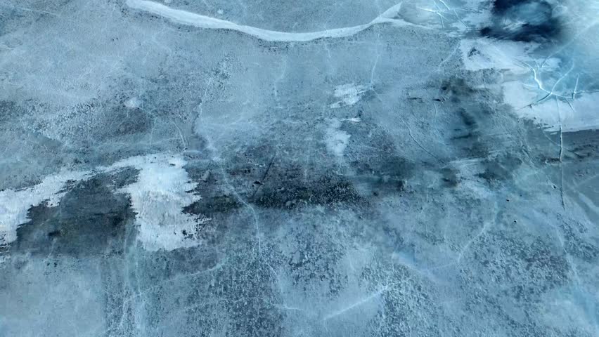 Graphic, top-down aerial shot of a deep fissure running through a sheet of thick blue ice during the spring thaw cycle.