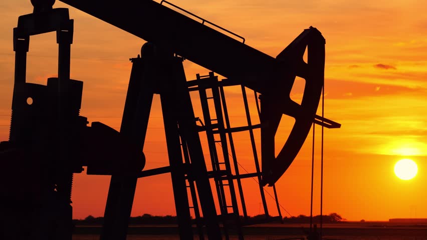 Silhouette of oil pump jack against a colorful beautiful sky with glowing clouds at sunset, high voltage electric pole in background