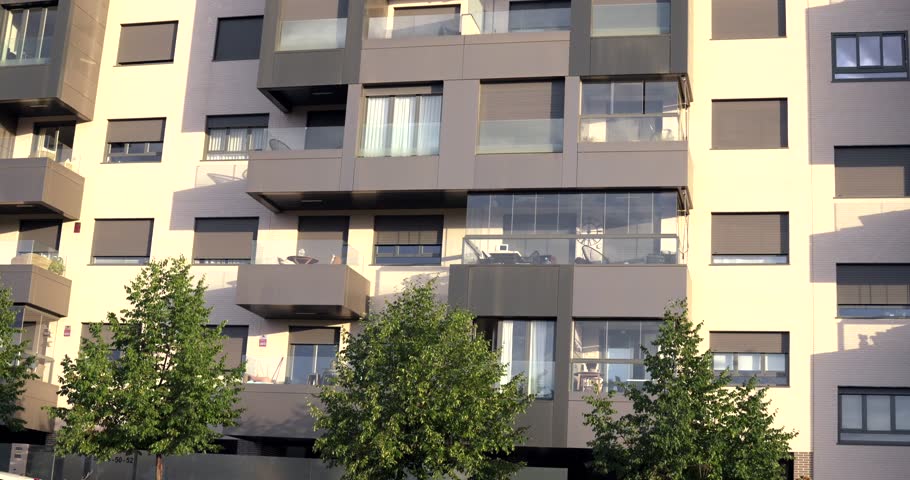 Close view of a modern residential apartment building featuring glass balconies, large windows and contemporary facade design. Urban housing architecture with trees in the foreground in Madrid, Spain.
