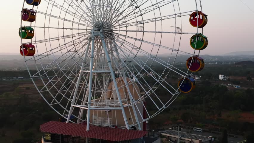 Ferris wheel with multicolored passenger gondolas stands against hazy sunset sky above rural terrain. Smooth forward tracking shot moves closer to the wheel structure, revealing detail of cabins.