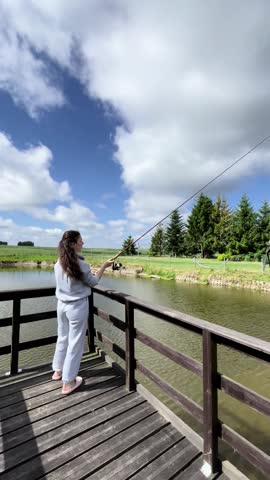 Young girl casts a fishing rod into a lake against the backdrop of a blue cloudy sky