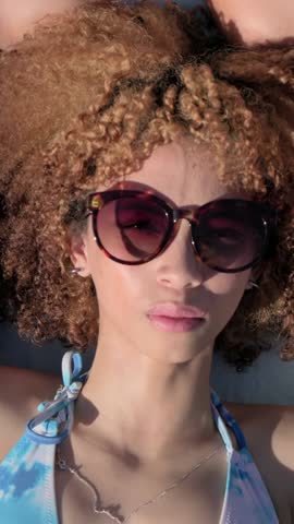 Vertical video: Camera widening revealing women sunbathing on beach mat with sunglasses and bikinis. Duo, towel, lifestyle, portrait, outdoors, sunlit, vertical video