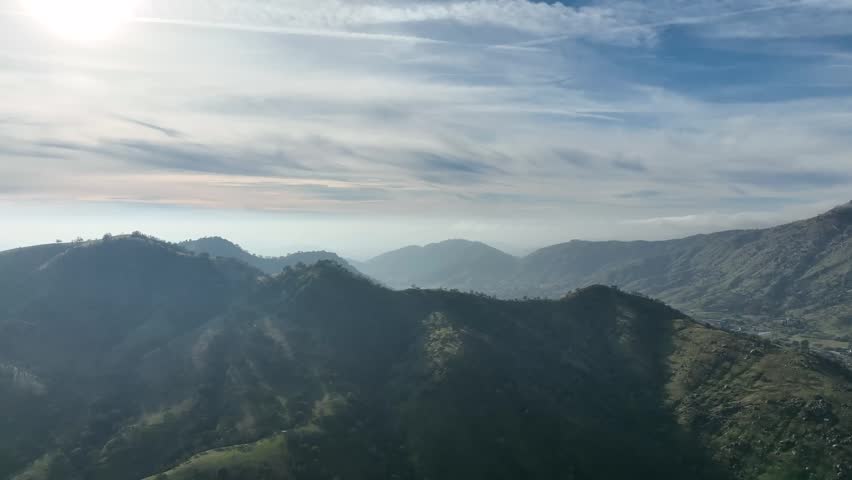 Aerial panoramic view of rolling mountain ridges and hazy valley under a bright cloudy sky in California
