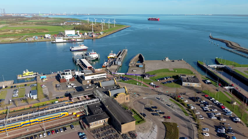 High-angle drone perspective showcasing a multi-modal transport hub in the coastal city of Vlissingen, Netherlands. The foreground highlights the train station tracks with iconic yellow and blue Dutch passenger trains, adjacent parking lots, and terminal buildings. The midground features the ferry port docks with passenger ships and vessels operating in the Westerschelde estuary. In the background, a large commercial cargo ship sails across the blue sea water toward the horizon, bordered by a green coastline equipped with industrial wind turbines under a clear blue sky.