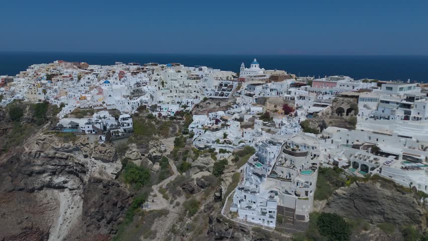 A panoramic view from a high vantage point shows the iconic village of Oia, with its densely clustered whitewashed houses, built along the steep volcanic cliffs of Santorini, Greece. The vibrant blue domes of the local churches stand out against a sea of white Cycladic architecture, which cascades down the dark, steep rock formations. Below, the deep blue waters of the Aegean Sea fill the caldera basin, dotted with small boats and yachts sailing close to the shore. The light blue horizon and the intense daylight create a striking, high-contrast scene that highlights the unique geographical location and charm of this popular Mediterranean destination.