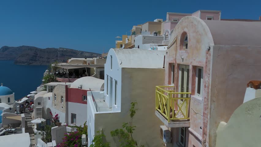A panoramic view from a high vantage point shows the iconic village of Oia, with its densely clustered whitewashed houses, built along the steep volcanic cliffs of Santorini, Greece. The vibrant blue domes of the local churches stand out against a sea of white Cycladic architecture, which cascades down the dark, steep rock formations. Below, the deep blue waters of the Aegean Sea fill the caldera basin, dotted with small boats and yachts sailing close to the shore. The light blue horizon and the intense daylight create a striking, high-contrast scene that highlights the unique geographical location and charm of this popular Mediterranean destination.