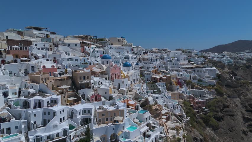 A panoramic view from a high vantage point shows the iconic village of Oia, with its densely clustered whitewashed houses, built along the steep volcanic cliffs of Santorini, Greece. The vibrant blue domes of the local churches stand out against a sea of white Cycladic architecture, which cascades down the dark, steep rock formations. Below, the deep blue waters of the Aegean Sea fill the caldera basin, dotted with small boats and yachts sailing close to the shore. The light blue horizon and the intense daylight create a striking, high-contrast scene that highlights the unique geographical location and charm of this popular Mediterranean destination.