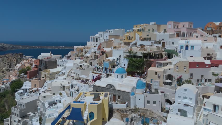A panoramic view from a high vantage point shows the iconic village of Oia, with its densely clustered whitewashed houses, built along the steep volcanic cliffs of Santorini, Greece. The vibrant blue domes of the local churches stand out against a sea of white Cycladic architecture, which cascades down the dark, steep rock formations. Below, the deep blue waters of the Aegean Sea fill the caldera basin, dotted with small boats and yachts sailing close to the shore. The light blue horizon and the intense daylight create a striking, high-contrast scene that highlights the unique geographical location and charm of this popular Mediterranean destination.