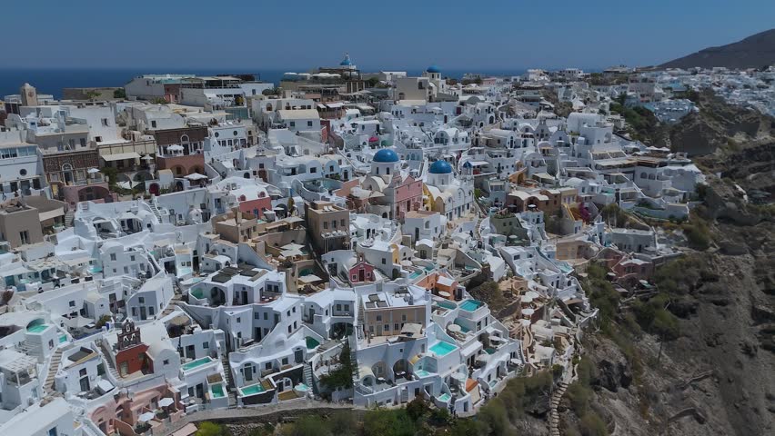 A panoramic view from a high vantage point shows the iconic village of Oia, with its densely clustered whitewashed houses, built along the steep volcanic cliffs of Santorini, Greece. The vibrant blue domes of the local churches stand out against a sea of white Cycladic architecture, which cascades down the dark, steep rock formations. Below, the deep blue waters of the Aegean Sea fill the caldera basin, dotted with small boats and yachts sailing close to the shore. The light blue horizon and the intense daylight create a striking, high-contrast scene that highlights the unique geographical location and charm of this popular Mediterranean destination.