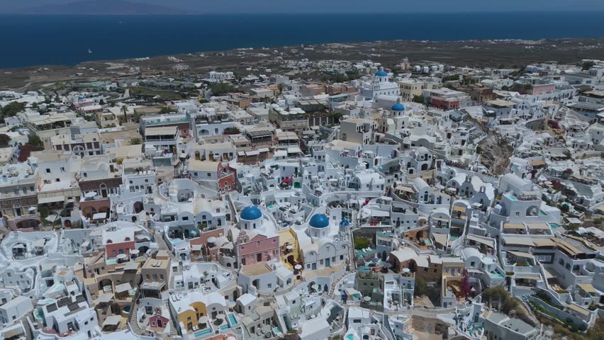A panoramic view from a high vantage point shows the iconic village of Oia, with its densely clustered whitewashed houses, built along the steep volcanic cliffs of Santorini, Greece. The vibrant blue domes of the local churches stand out against a sea of white Cycladic architecture, which cascades down the dark, steep rock formations. Below, the deep blue waters of the Aegean Sea fill the caldera basin, dotted with small boats and yachts sailing close to the shore. The light blue horizon and the intense daylight create a striking, high-contrast scene that highlights the unique geographical location and charm of this popular Mediterranean destination.