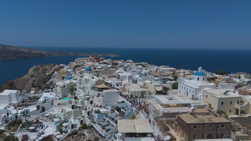 A panoramic view from a high vantage point shows the iconic village of Oia, with its densely clustered whitewashed houses, built along the steep volcanic cliffs of Santorini, Greece. The vibrant blue domes of the local churches stand out against a sea of white Cycladic architecture, which cascades down the dark, steep rock formations. Below, the deep blue waters of the Aegean Sea fill the caldera basin, dotted with small boats and yachts sailing close to the shore. The light blue horizon and the intense daylight create a striking, high-contrast scene that highlights the unique geographical location and charm of this popular Mediterranean destination.