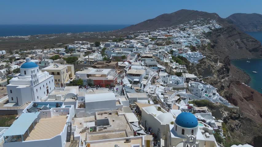 A panoramic view from a high vantage point shows the iconic village of Oia, with its densely clustered whitewashed houses, built along the steep volcanic cliffs of Santorini, Greece. The vibrant blue domes of the local churches stand out against a sea of white Cycladic architecture, which cascades down the dark, steep rock formations. Below, the deep blue waters of the Aegean Sea fill the caldera basin, dotted with small boats and yachts sailing close to the shore. The light blue horizon and the intense daylight create a striking, high-contrast scene that highlights the unique geographical location and charm of this popular Mediterranean destination.