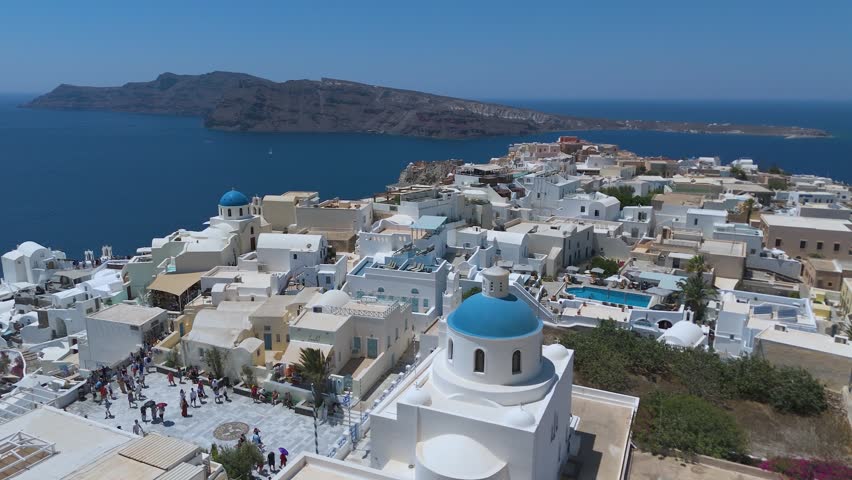 A panoramic view from a high vantage point shows the iconic village of Oia, with its densely clustered whitewashed houses, built along the steep volcanic cliffs of Santorini, Greece. The vibrant blue domes of the local churches stand out against a sea of white Cycladic architecture, which cascades down the dark, steep rock formations. Below, the deep blue waters of the Aegean Sea fill the caldera basin, dotted with small boats and yachts sailing close to the shore. The light blue horizon and the intense daylight create a striking, high-contrast scene that highlights the unique geographical location and charm of this popular Mediterranean destination.