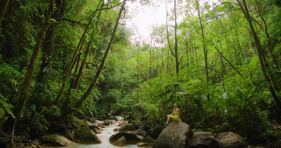 Tourist woman sitting on a large rock by a jungle river in Costa Rica during journey through wold scenic landscapes