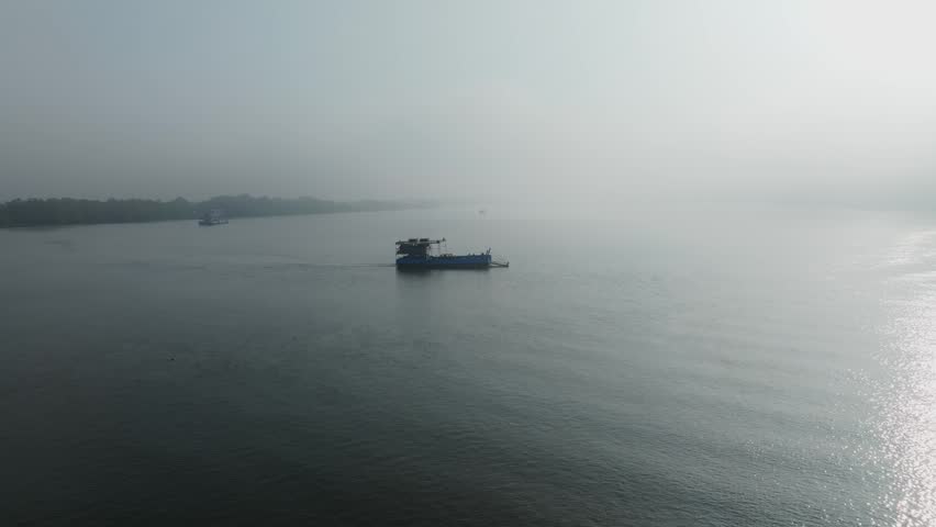 Ferry boat crossing Mandovi river through calm water in early morning fog. Aerial view captures misty atmosphere with forested shoreline and soft dawn light during misty foggy sunrise.