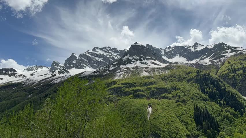 Snow-covered peaks tower over vibrant green hills under a clear blue sky filled with white clouds, showcasing a stunning mountain landscape