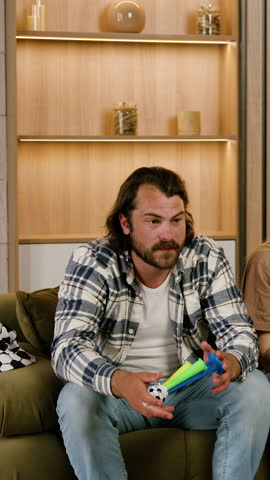 Male gamer with dark hair in plaid shirt reacts intensely while playing video game on couch, raising arms in excitement in a cozy living room setting