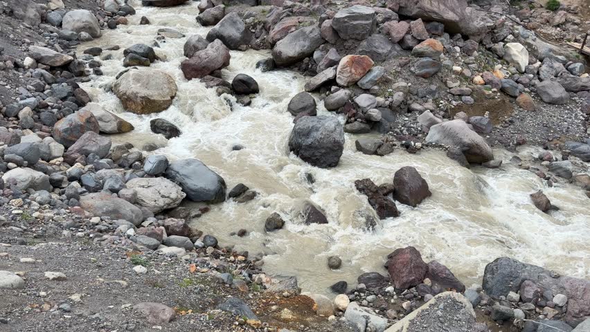 River water flows rapidly over stones, with a wooden bridge crossing the stream, highlighting the natural beauty of the mountainous terrain
