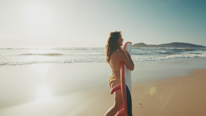 Sexy woman surfer in sensual bikini walks on the sandy beach with surfing board on sunrise