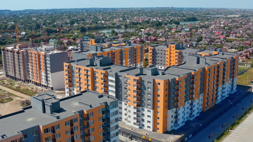 Dynamic aerial view reveals modern residential apartment complex featuring bright orange and gray buildings, surrounded by green landscape, with construction site in background, on sunny day.