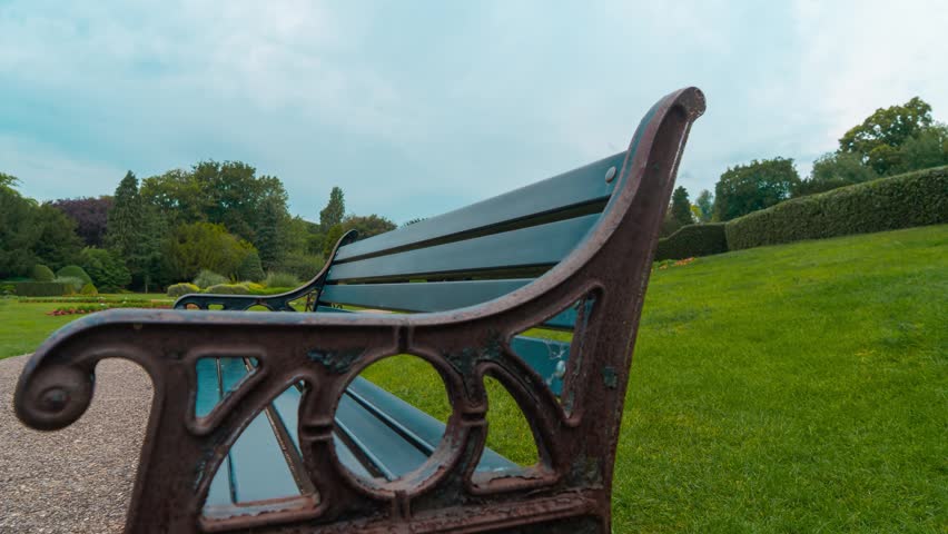 Vintage park bench overlooking green lawn during smooth slider timelapse. Fast moving clouds creating dramatic evening sky above peaceful public garden landscape. Cinematic outdoor scenery
