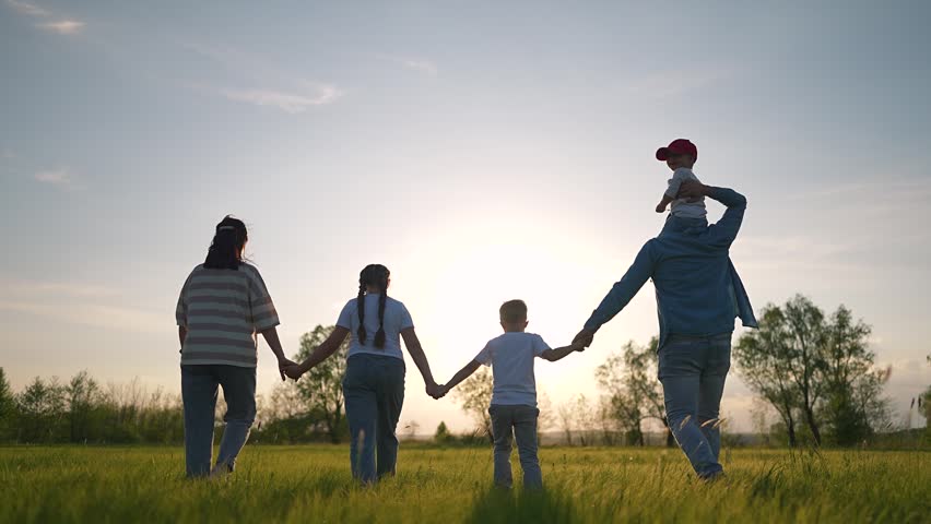Family holding hands walking through meadow. Boy runs ahead, father holds child, girl walks near mother. Sunset light on meadow. Family, boy, child move across field. Family bonds strong during walk