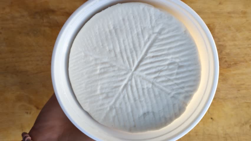 Top view of a person unmolding a wheel of fresh white goat cheese from a plastic mold container onto a rustic wooden board, traditional cheese making and gourmet artisan food production