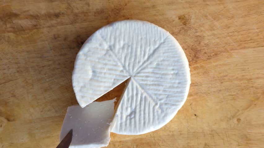 Top view of a person cutting a wheel of traditional white goat cheese on a rustic wooden board, gourmet food preparation and artisan cheese making concept in a Mediterranean kitchen