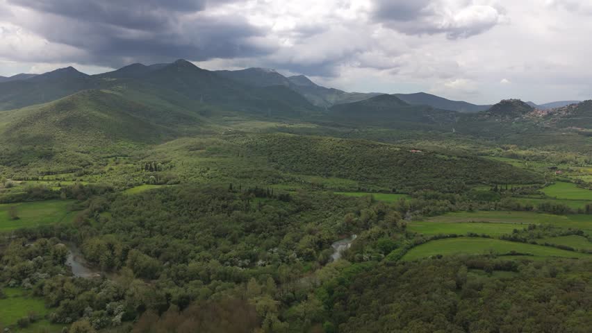 Wide aerial drone footage of a lush green mountain valley in the Peloponnese, Greece. The scene shows olive groves, forested hills, green fields, a winding river, distant villages on the slopes and layered mountain ranges under dramatic storm clouds. Soft sunlight breaks through the clouds and creates patches of light and shadow across the landscape. This footage works well for travel, nature, Mediterranean scenery, mountain valley backgrounds, rural landscapes, spring greenery, aerial establishing shots, environmental projects and peaceful outdoor visuals.