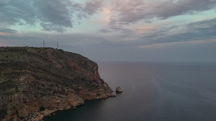 Stunning 4K aerial drone footage of Cabo de San Antonio capturing the dramatic rocky Mediterranean headland at dusk. Steep rocky cliffs with communications tower installation rise majestically above calm turquoise waters. Isolated rock formations and sea stacks frame the pristine coastline beneath soft pastel sky with gentle clouds. The serene seascape reflects the tranquility of this rugged Costa Blanca landmark. Perfect for travel, nature, and Mediterranean coastal destination content projects showcasing authentic landscape beauty.