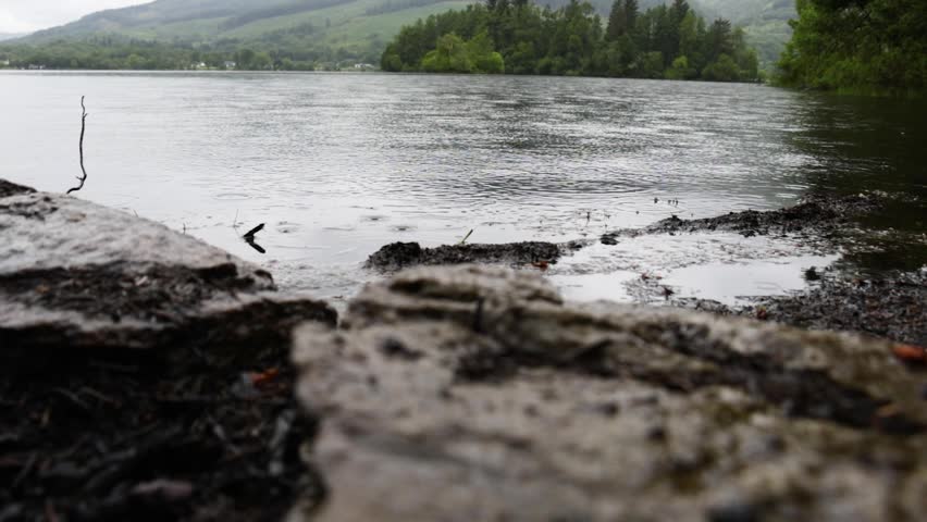 Rain on Loch Ard, Scotland, seen in a static close shot as droplets dimple the lake surface by a rocky shore with dark clouds and misty hills in the background.