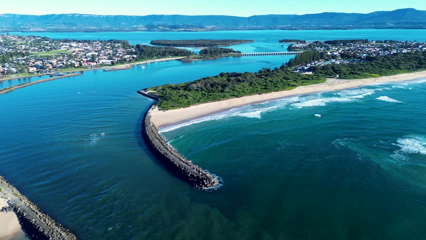 Drone aerial landscape of ocean waves breaking along Windang Beach beside the passage break wall inlet with the town bridge and Lake Illawarra in the background near Wollongong South Coast Australia