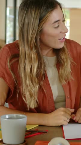 Vertical video: Responding to right-side voice, woman in rust top turning at table, tapping notepad. Conversation, kitchen, dining, window, sunlit, natural, vertical video