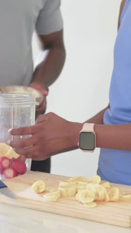 Vertical video: Mixing yogurt, man in tee reaching spoon at kitchen counter with blender for shake. Couple, fruit, banana, pineapple, blueberries, creaminess, vertical video