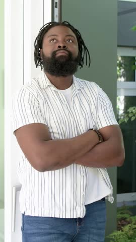 Vertical video: Panning camera, man shifting gaze and smiling in door, wrist bracelet, copy space. Adult, person, porch, entryway, daylight, greenery, vertical video