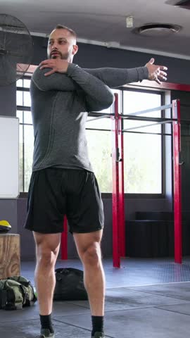Vertical video: Stretching man in grey top doing upper-body stretches at gym before workout, rig. Mobility, crossbody, triceps, warmup, athletic, daylight, vertical video