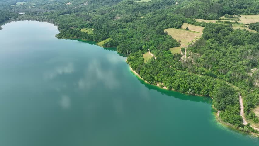 Turquoise water of Butoniga reservoir reflects white clouds in Istria region. Drone flies over artificial lake surrounded by dense green forests and rolling hills near Motovun.