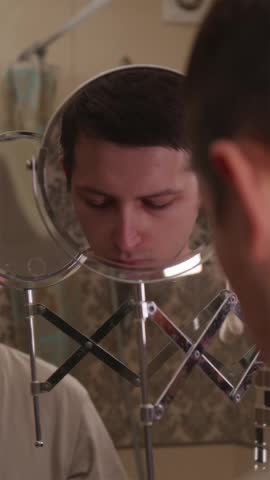Young gentleman using a precise electric foil shaver machine to remove stubble from his cheek while looking into a cosmetic mirror.