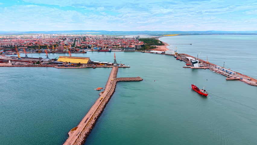 Aerial view of cargo ship entering industrial port channel. Red cargo vessel navigating into port harbor entrance between breakwaters with cranes and docks visible.