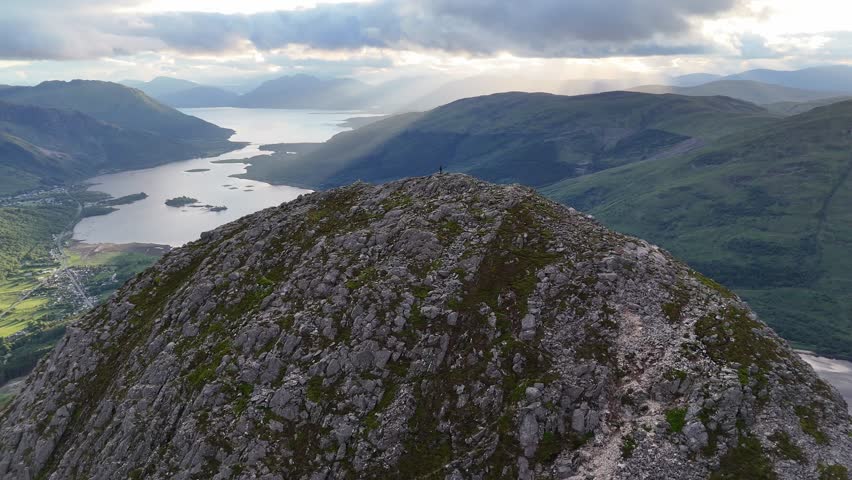 This aerial drone footage is a tracking shot that pushes forward towards a lone person standing on the rugged rocky peak of Pap of Glencoe in Scotland UK.