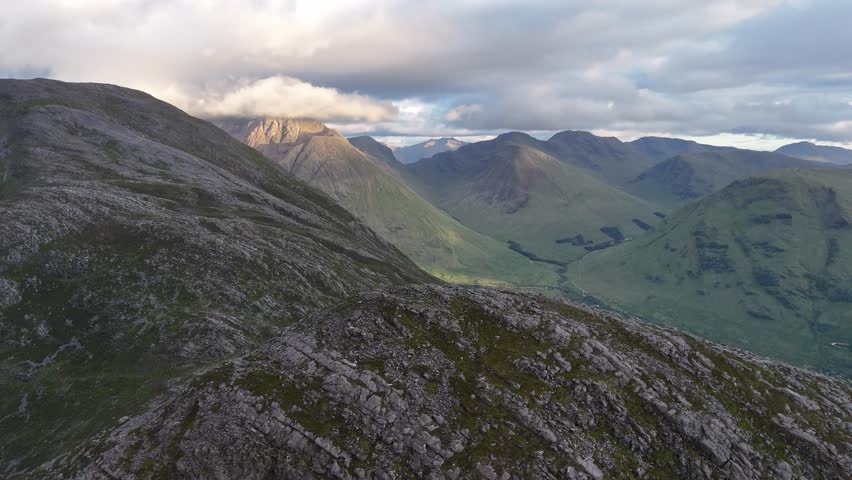 Dramatic drone tracking shot over the rugged, rocky summit of Pap of Glencoe, revealing a breathtaking vista of a deep valley, Loch Leven, and surrounding mountains in the Scottish Highlands, UK.